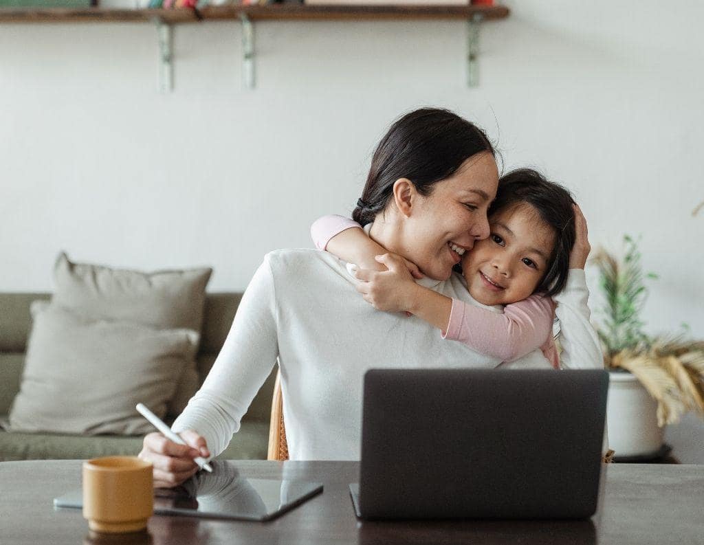 Eine Mutter sitzt am Schreibtisch und arbeitet am Laptop, während sie ihr Kind liebevoll umarmt – ein Beispiel für die Herausforderung, Beruf und Privatleben in der Work-Life-Balance zu vereinen.
